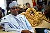 Former Nigerian President Muhammadu Buhari smiling as he sits on a chair with a gold edge and wears a blue outfit.