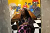 An image of the designer sitting on a chair that\u2019s placed on a checkered floor and there\u2019s a vibrant art piece behind her.