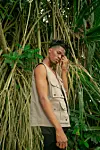 Kwesi Arthur stands in front of bamboo wearing a beige vest, black pants