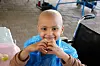 A young bald child, wearing golden hoop earrings and a blue plastic overall, is smiling into the camera and forming a heart with their hands.