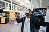 A man wearing virtual reality goggles holds up a control. There are various video games on tv's in the background at a convention center.