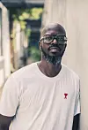 South African DJ Black Coffee stands, wearing a white tee shirt and clear glasses.