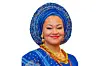 Senator Natasha Akpoti in a beaded blue traditional Nigerian outfit with a matching blue head wrap (gele), gold earrings, colorful necklaces, and red lipstick, smiling at the camera.