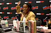 Tomi Adeyemi speaks energetically at New York Comic Con, seated behind a table with fantasy novels and microphones, she is smiling, and her hair is in locks.