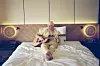 Salif Keita, dressed in yellow traditional clothing, sits barefoot on a neatly made bed in a softly lit room, playing an acoustic guitar with a thoughtful expression.