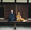 Two men sit peacefully on a wooden veranda; one in a traditional Japanese robe seated in a meditative pose, and the other, Salif Keita, in mustard-yellow West African attire with bare feet, both exuding calm against the backdrop of wooden sliding panels.