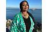 Sudanese American radio journalist Hana Baba stands in front of the sea, wearing a green kimono, matching green jewelry, and a black hijab. She is smiling into the camera.