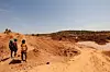 A group of men stand on reddish-brown earth at an informal mining site, surrounded by mounds of excavated soil and a water-filled pit. In the background, more miners can be seen working under a clear blue sky, with sparse vegetation on the horizon.