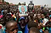 A large crowd of Ugandan supporters gather at a political rally, with several people holding up campaign posters of opposition leader Dr. Kizza Besigye. The mood is energetic, with people cheering, raising peace signs, and blowing whistles.