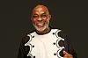 Richard Mofe-Damijo making a dance pose and smiling in a black outfit with a bold white patterned design.