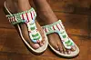 The feet of a model wearing beaded sandals with green, red, and white patterns stands on a wooden floor.