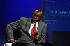 Strive Masiyiwa, sits on stage, wearing a grey suit and red tie, holding a microphone. He appears focused and thoughtful, with a cup labeled “U.S.-Africa Business Forum” placed in front of him. The background is lit in deep blue tones, creating a formal and professional atmosphere.