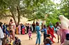 A street filled with children and women in colorful clothes, some are holding containers in which they will pick up the day\u2019s meal.