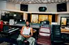 A photo of GuiltyBeatz sitting in a studio wearing white singlet and silver baggy pants.