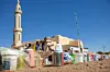 Containers and Tupperware are lined under blue skies outside a mosque. In the distance, young men are sitting by the mosque and looking towards the camera while older men are entering it.