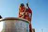 Under a cloudless blue sky, an elderly woman wrapped in an orange toub is pounding dough in a pot on the ground with a wooden stick.