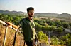 Omer Al Tijani is standing on a hill, wearing a green t-shirt and smiling. Behind him are lush trees and mountains.