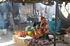 A woman wearing a toub with orange and purple flowers sits in a makeshift stall at a food market, selling fresh vegetables.