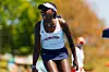 Angella Okutoyi wearing an Auburn University tennis uniform, shouting in celebration during a match, with a blurred outdoor background and a visible official in the distance.