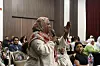 A woman wearing a beige and pink hijab is addressing people in a conference room, speaking into a microphone and gesticulating with a lifted arm.