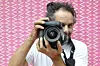 Hassan Hajjaj is holding a camera in front of his face, standing in front of a pink patterned wall, and wearing a white t-shirt.