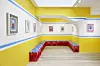 A nook filled with simple red seats in a gallery with blue, white and yellow walls hung with Hajjaj's artwork.