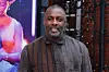 Idris Elba wearing a long, striped shirt with shades of brown and gray, posing outdoors and smiling warmly.