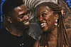 A couple shares a warm smile; the man wears a black shirt and a necklace with a gold pendant, while the woman, with long braids with colorful beads, wears elegant earrings and a gold necklace, set against a vibrant, artistic background.