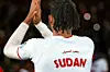A football player is seen from behind, wearing a white jersey with red accents that reads “SUDAN” and the number 11. Arabic text is written above the country’s name. The player is clapping, under bright stadium lights, with blurred spectators in the background.