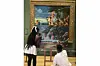 Two young artists work on a striking modern painting of a multi-headed serpent attacking warriors, placed on an easel in front of a classical Renaissance artwork in a museum, blending old and new artistic styles in one powerful scene.