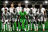 The Sudan national football team posing for a group photo on the field before a match. The players are dressed in white jerseys with red detailing, except for the goalkeeper who is wearing a bright green kit. The team is arranged in two rows, with the front row kneeling and the back row standing.