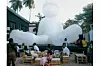 A group of people gather around wooden pallet seating in an outdoor courtyard, in front of a massive white inflatable figure with goggles, surrounded by palm trees and buildings.
