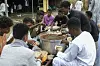 Group of young Sudanese men sharing a communal meal from a large pot, serving food into plastic bags in an outdoor community setting.