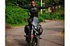 Ebaide Udoh beams with joy as she stands on a motorcycle, arms raised in victory, wearing full riding gear and sunglasses, surrounded by lush greenery on a quiet road.