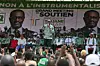 A crowd gathers at a political rally in Côte d’Ivoire in support of Tidjane Thiam’s 2025 presidential campaign. Thiam’s image dominates a large campaign banner in the background, while a speaker addresses supporters from the stage, surrounded by party members wearing coordinated green and white outfits