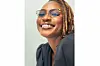 A smiling Ebaide Udoh with light brown locks and cat-eye glasses looks directly at the camera. Her glossy lips and clear skin catch the light. She wears a dark top with puffed sleeves and exudes joy and confidence.