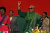 Namibia's first female president, Netumbo Nandi-Ndaitwah stands with her fist in the air. She is smiling and wears a green outfit and sunglasses. There are three women next to hear and a bouquet of flowers in front of her.
