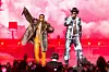 Wizkid and Burna Boy perform on stage. Wizkid is in a fur coat and orange leather pants, Burna Boy is in a black cowboy hat and graphic suit, against a backdrop of pink clouds and red lighting.