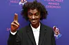 A photo of K'naan, wearing a black suit and smiling as he raises his hand in a peace sign gesture at the Recording Academy's Special Merit Awards Ceremony at the Wilshire Ebell Theatre in Los Angeles, California, on February 3, 2024.