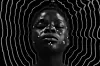 A black and white artistic portrait of a person with dark skin, eyes closed, and adorned with small white facial markings. White lines ripple around the person's head to make frequency-like waves around the person's head, creating a halo-like effect against the dark background.