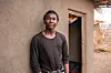 Espoir Kahitani, a young man in a black shirt, stands beside a house in Dzaleka Refugee Camp.