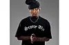 Xduppy stands against a plain gray background, gazing directly into the camera with a serious expression. He wears a black oversized T-shirt that reads “Scotty Uzi” in bold gothic script and a slouchy black beanie tilted to one side, partially covering his bleached-tipped locks. His hands are clasped together in front of him, and he accessorizes with a silver chain necklace and matching bracelets.