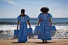 Two girls, one with curly afro hair, the other with two braids, standing at the beach, facing the sea, wearing blue and white checkered dresses and carrying bags of the same textile.
