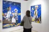 A woman is standing in front of two blue paintings that show abstract people in traditional clothes.