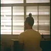 A person with braided hair sits with their back facing the camera, gazing out of a window with frosted glass.