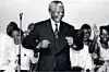 A photo of Nelson Mandela wearing a suit while smiling as he does his famous “Madiba shuffle” which involves forming a double fist and raising both arms in a curved position while moving the body slightly.