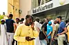 A concerned-looking man in a yellow jalabiya is holding his phone outside a Bank of Khartoum branch where several other men appear to be waiting with him.