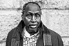 Late Kenyan author Ngũgĩ wa Thiong’o stands in front of a stone wall, wearing a patterned shirt and a dark coat with a scarf draped over his shoulders.
