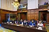 Adelegation from Sudan seated at a long table inside an ornate chamber at the ICJ. The architecture features carved wood paneling, a grand chandelier, and a gallery with observers above.