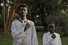 A man wearing a white jalabiya and a beige tie stands next to a boy wearing a jalabiya. They are in the middle of a forest, solemnly holding their right fists over their chest as one does when listening to a national anthem.